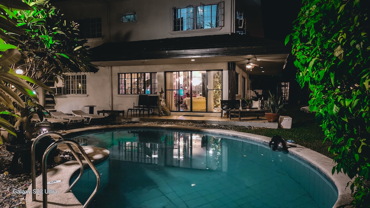 A serene pool area is surrounded by lush greenery, featuring a well-maintained swimming pool with a gentle curve. Large glass doors open to a spacious living area, providing an inviting connection between indoor and outdoor spaces, complemented by ambient lighting.