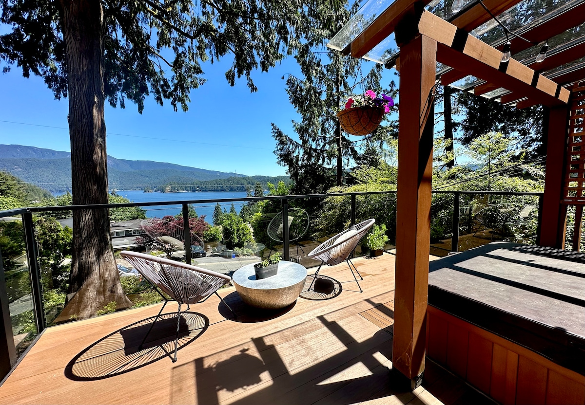 A private deck overlooks the water and surrounding landscape. Two comfortable chairs are positioned beside a circular table, with a hot tub visible at the far end. A pergola provides partial shade, and hanging plants add a touch of greenery to the serene setting.