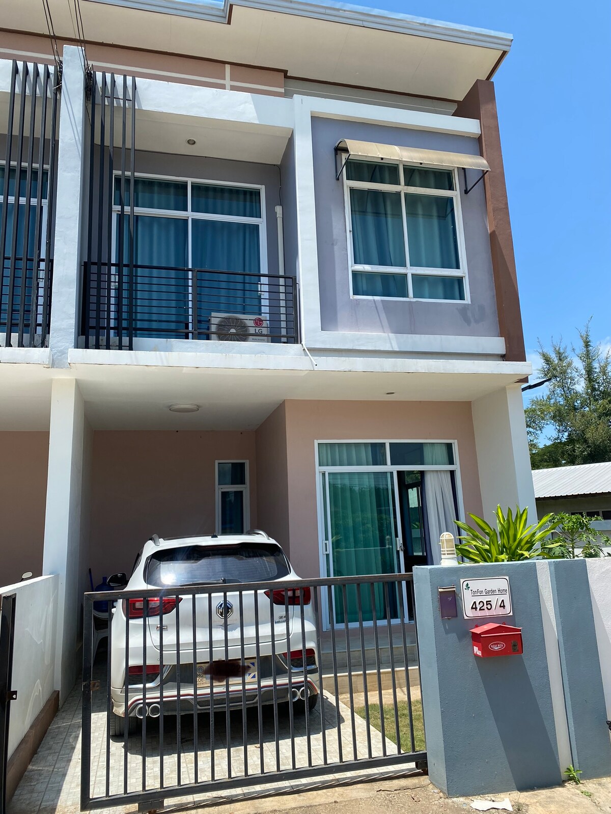 A modern townhome exterior is showcased with a fenced driveway that accommodates a parked vehicle. Large windows allow natural light to illuminate the entrance area. The facade features a combination of soft colors, and small plants are visible near the entrance, enhancing the overall appeal.