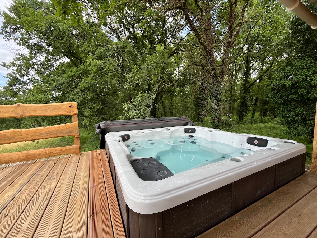 A hot tub with a white shell is situated on a wooden deck, surrounded by lush green trees. The clear water invites relaxation, complemented by a wooden railing that offers a sense of privacy and integration with nature.