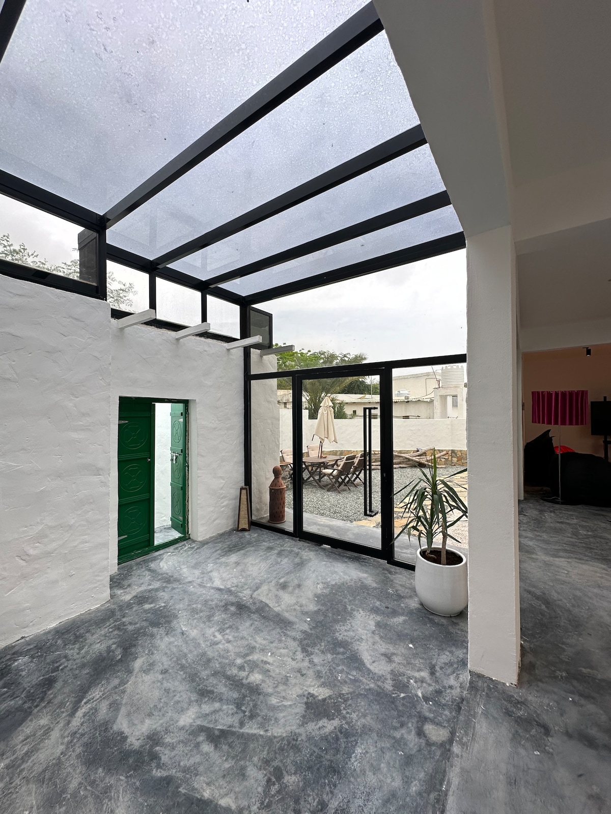A bright entryway features a green door framed by large glass windows, allowing natural light to fill the space. A potted plant adds a touch of greenery to the corner, while views of an outdoor seating area and patio are visible through the glass walls.