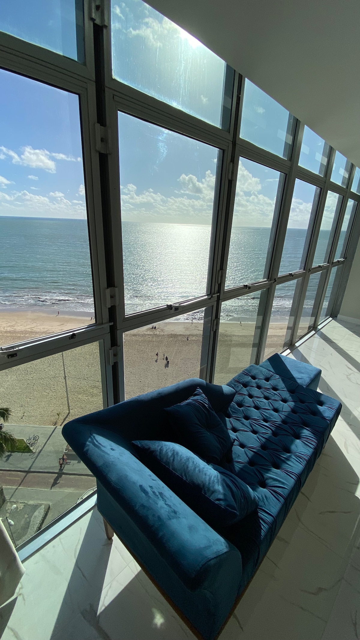 A plush blue sofa is positioned near large floor-to-ceiling windows, offering a view of the ocean and beach. Soft sunlight reflects off the water, creating a serene atmosphere. The spacious room is filled with natural light, enhancing the inviting space.