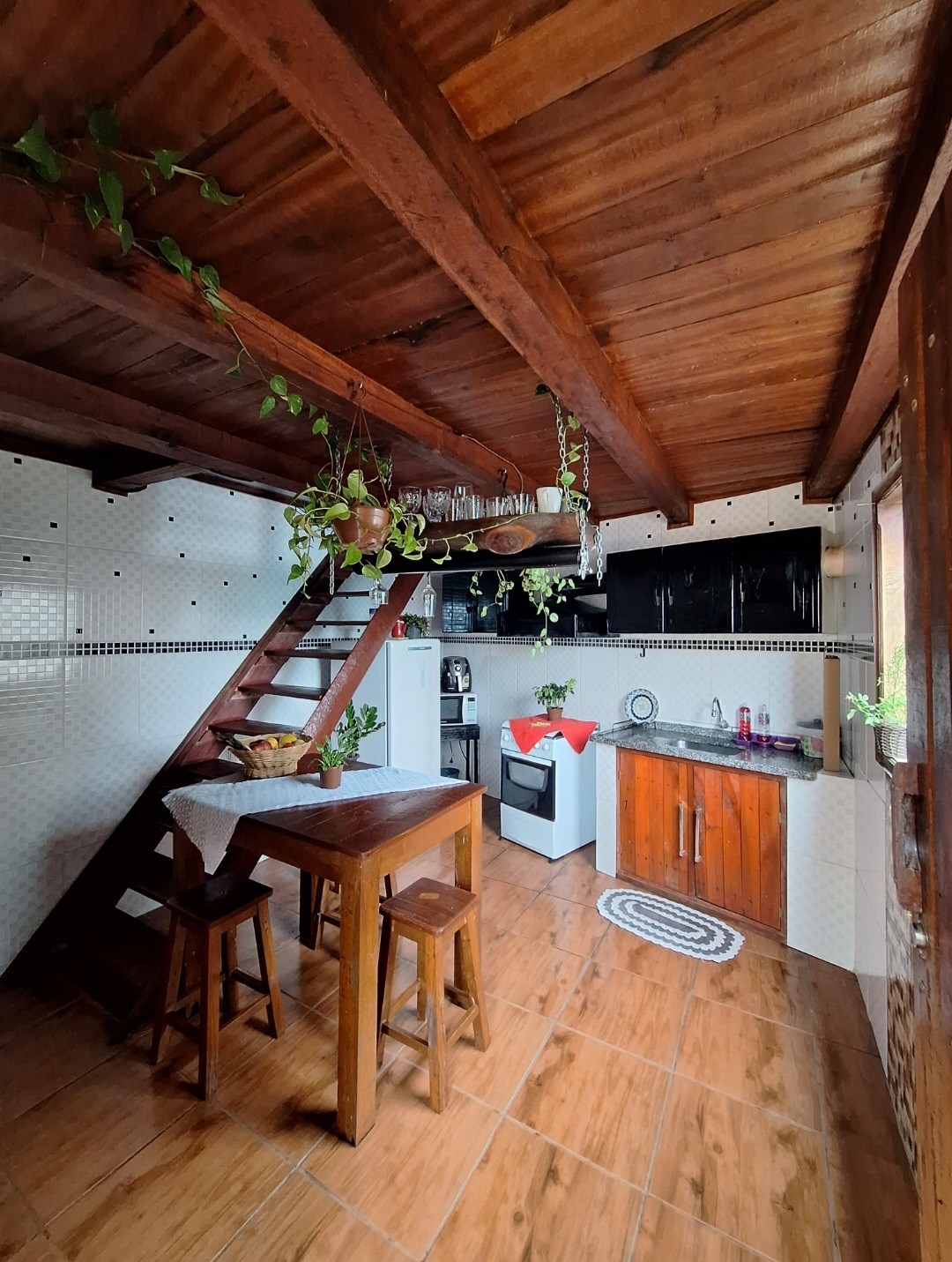 An open kitchen showcases wooden beams and tiled walls. A small dining table with two stools sits centrally, surrounded by greenery. A staircase leads to the upper level, while appliances, including a stove and refrigerator, are neatly arranged along the walls.