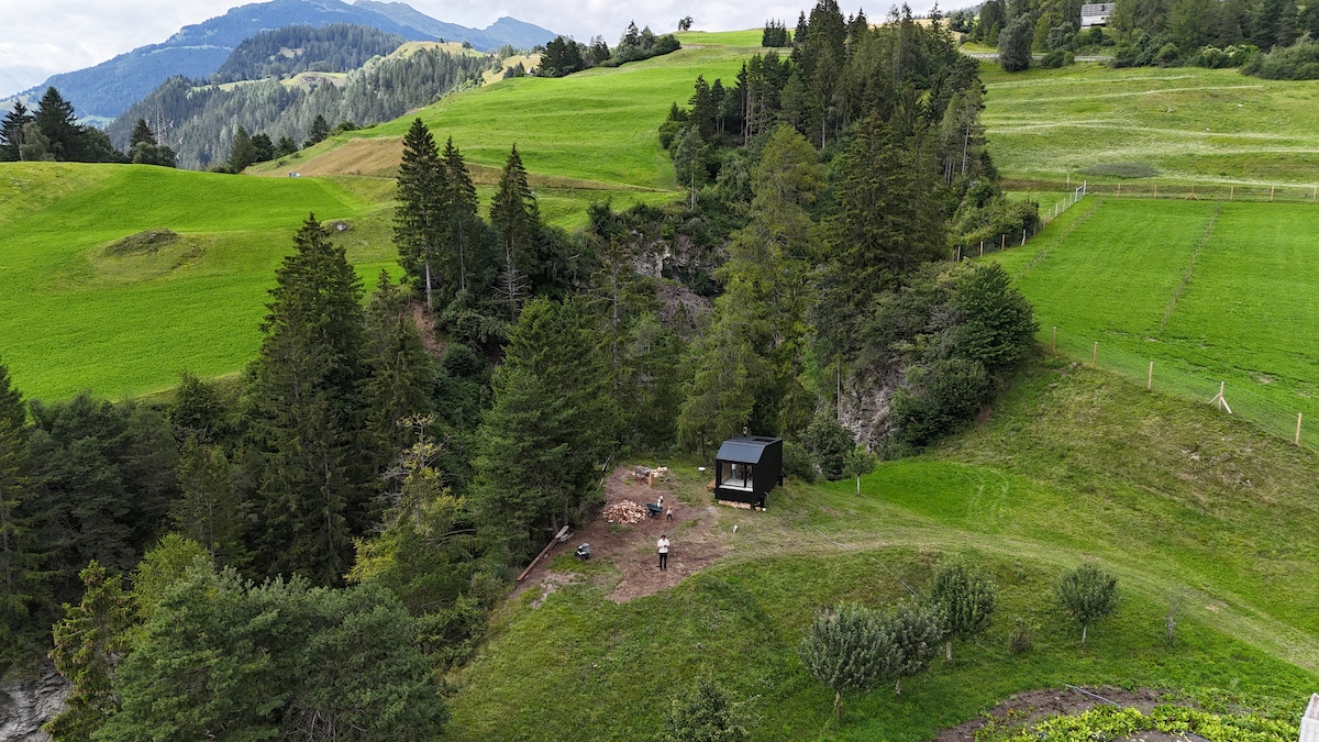 A secluded cabin is nestled within lush greenery, surrounded by rolling hills and trees. The landscape features vibrant grass and a few scattered bushes. A fire pit and chairs can be seen near the cabin, inviting relaxation in a tranquil outdoor setting.