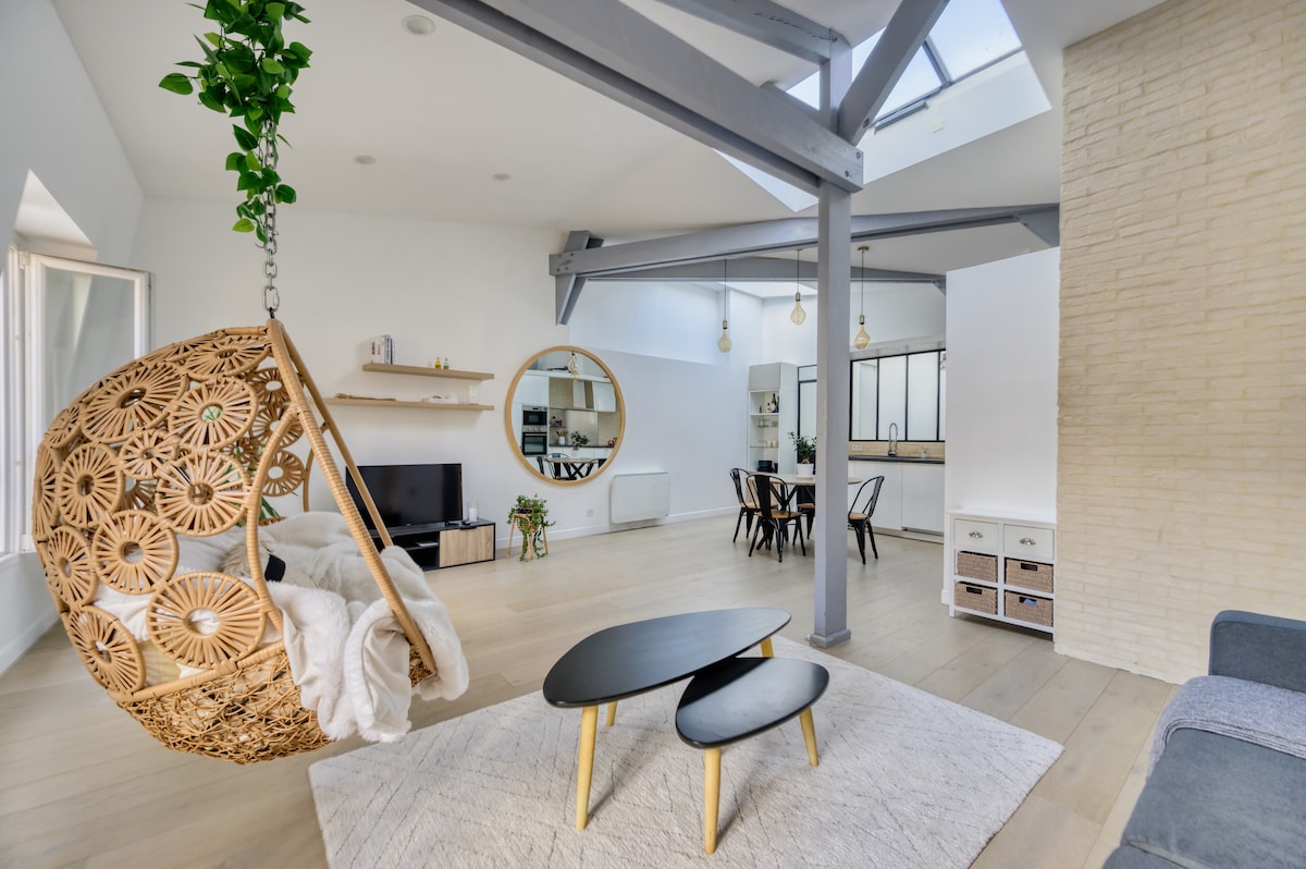 A spacious living area is highlighted by a woven hanging chair and a round mirror. A minimalist coffee table sits atop a light area rug, with wood flooring extending throughout. The open kitchen and dining space are visible in the background, defined by modern furnishings.