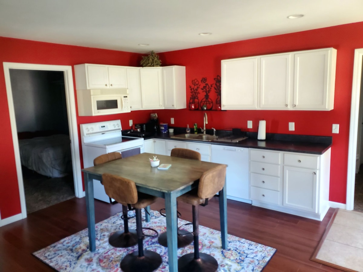 A kitchen features white cabinetry and a black countertop, contrasted by a bold red accent wall. A wooden table with four brown chairs is positioned in the center, while essential appliances and cooking items are organized on the countertops.