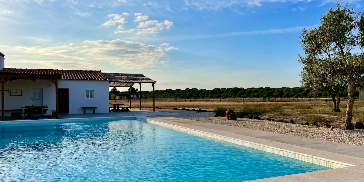 A large salt-water pool is positioned in the foreground, reflecting the clear sky and surrounding landscape. A shaded seating area is located beside the pool, with a simple white building visible in the background, framed by trees and open prairie.