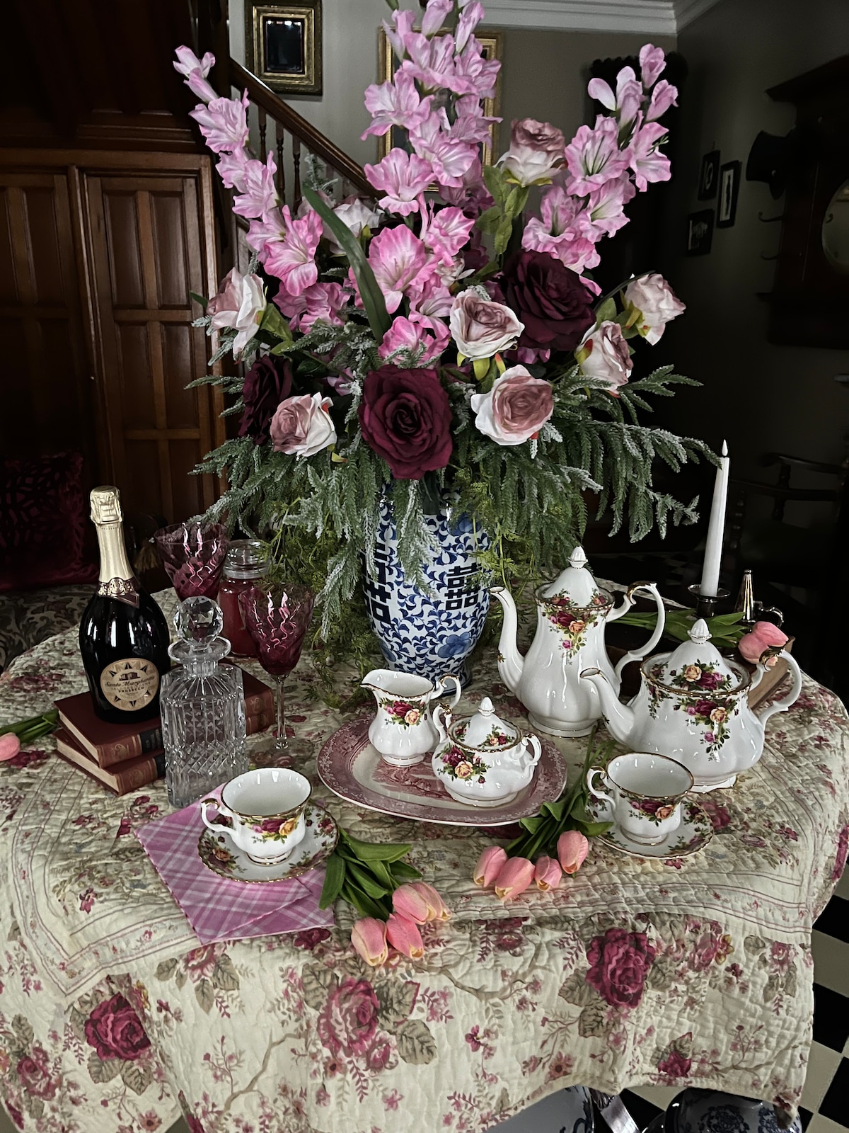 A decorative table setting features an ornate floral tablecloth adorned with vintage-style china, including a teapot and cups. Fresh pink tulips and an extravagant floral arrangement add vibrancy. Other elements include a crystal decanter, candle holder, and a bottle of sparkling wine, enhancing the elegant presentation.