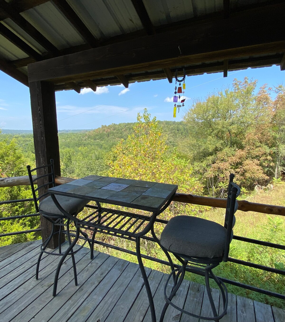 A shaded outdoor seating area features a small square table accompanied by two black metal chairs. Lush green hills are visible in the background, under a clear blue sky. A wind chime adds a decorative touch to the rustic surroundings.