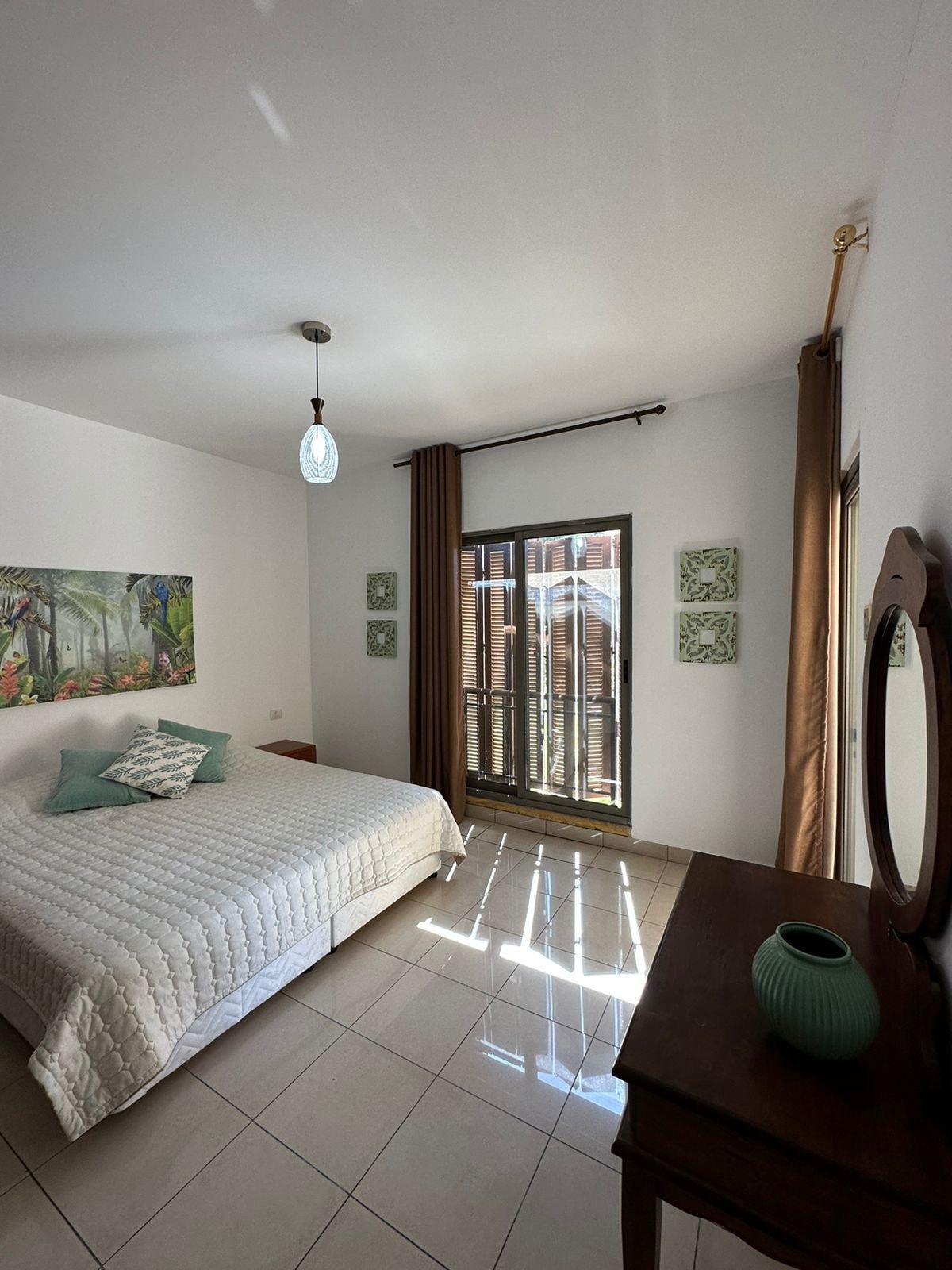 A cozy bedroom features a spacious bed with a white quilted coverlet, complemented by soft pillows. Artwork adorns the wall, and natural light filters through the large window, which is framed by brown curtains. A wooden dresser and a decorative vase complete the serene setting.