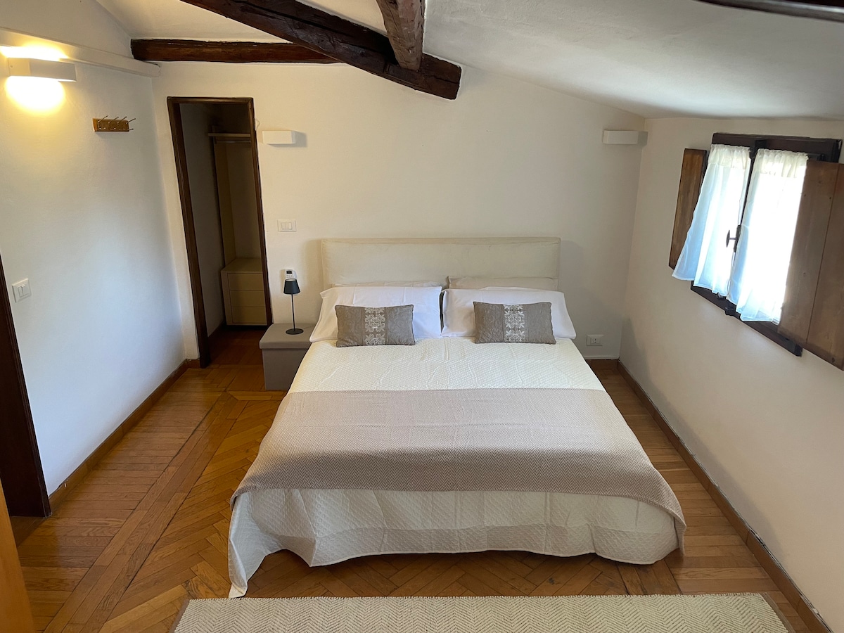 A cozy bedroom features a spacious bed with a light-colored blanket, complemented by soft pillows. Natural light filters through the window, which is adorned with light curtains. Exposed wooden beams add character, while a simple bedside lamp and storage area are visible.