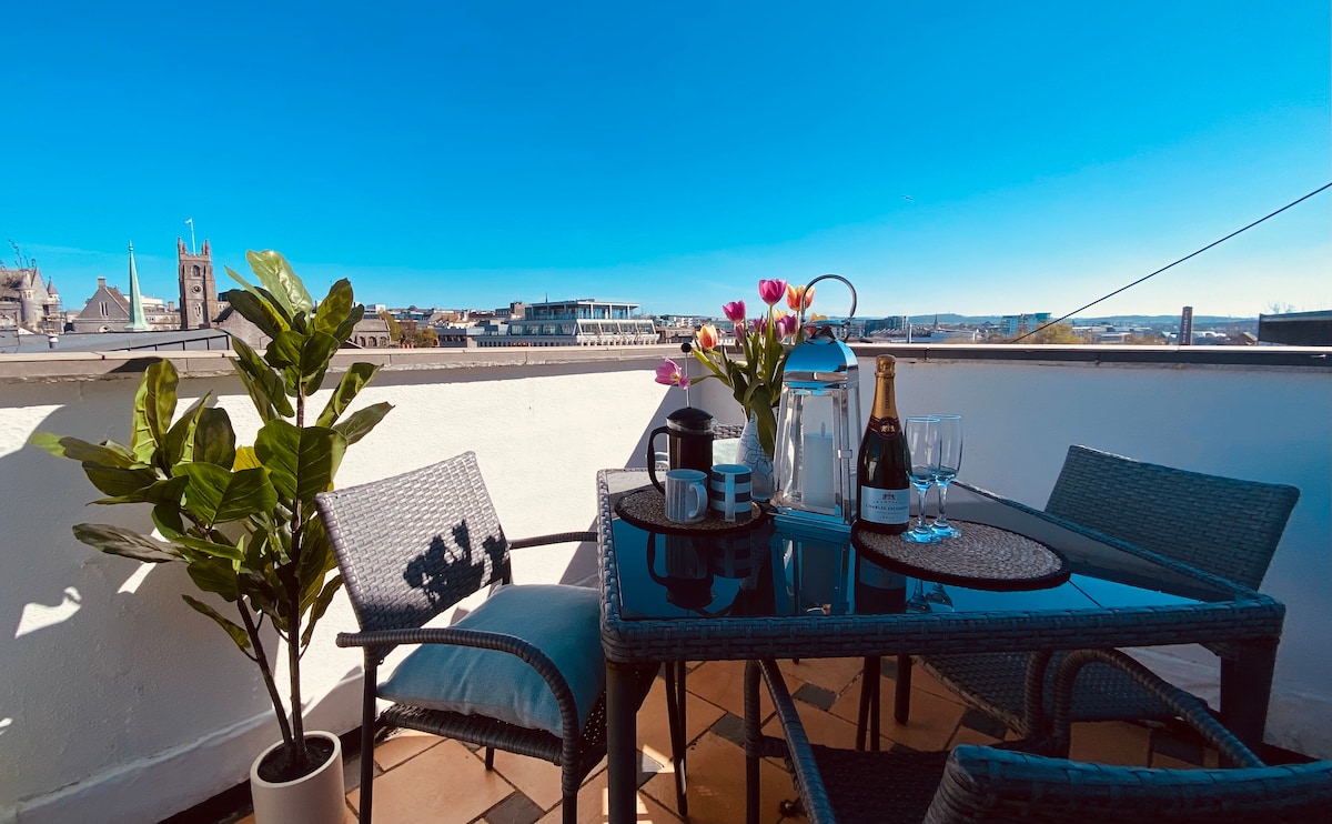 A roof terrace is presented with a table and chairs, featuring a small vase of flowers. Two glasses and a bottle of champagne rest on the table, set against a backdrop of clear blue skies and views of Plymouth's skyline.