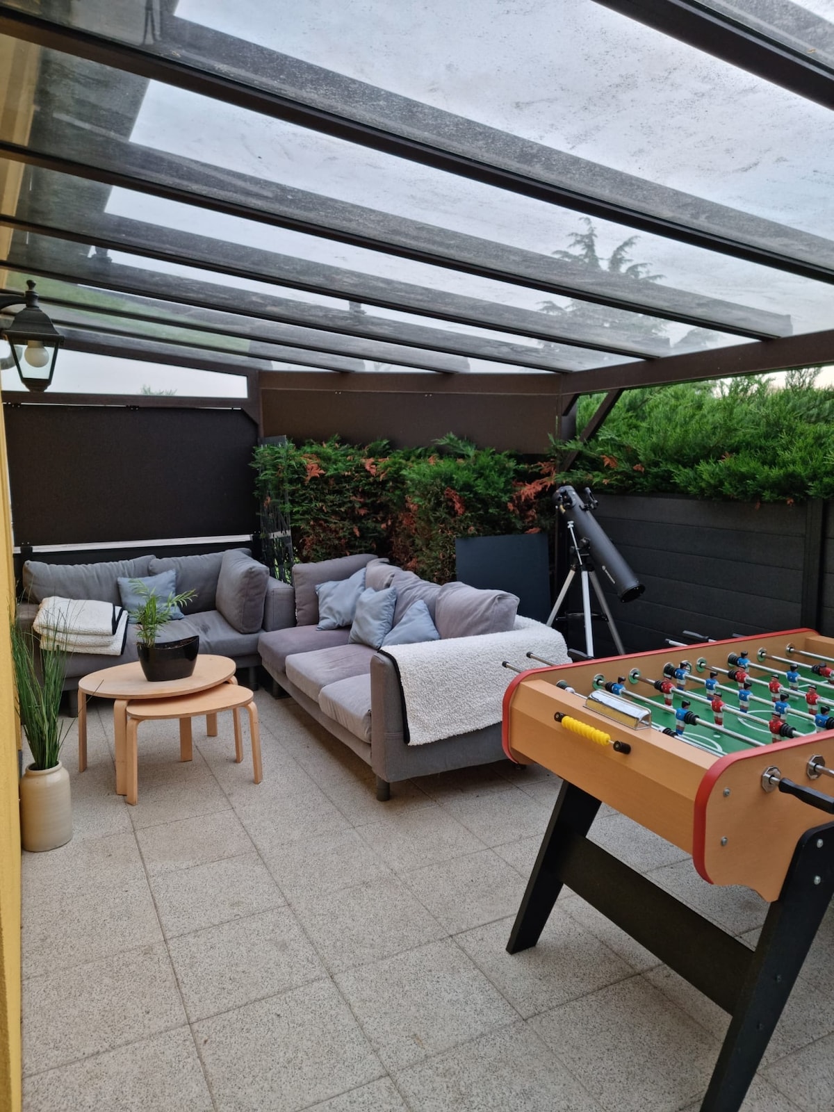 A covered outdoor lounging area is depicted, featuring a comfortable seating arrangement with soft cushions and a throw blanket. A small table accompanies the seating, while a foosball table is positioned nearby. Potted plants add greenery to the space, with a telescope visible in the background.