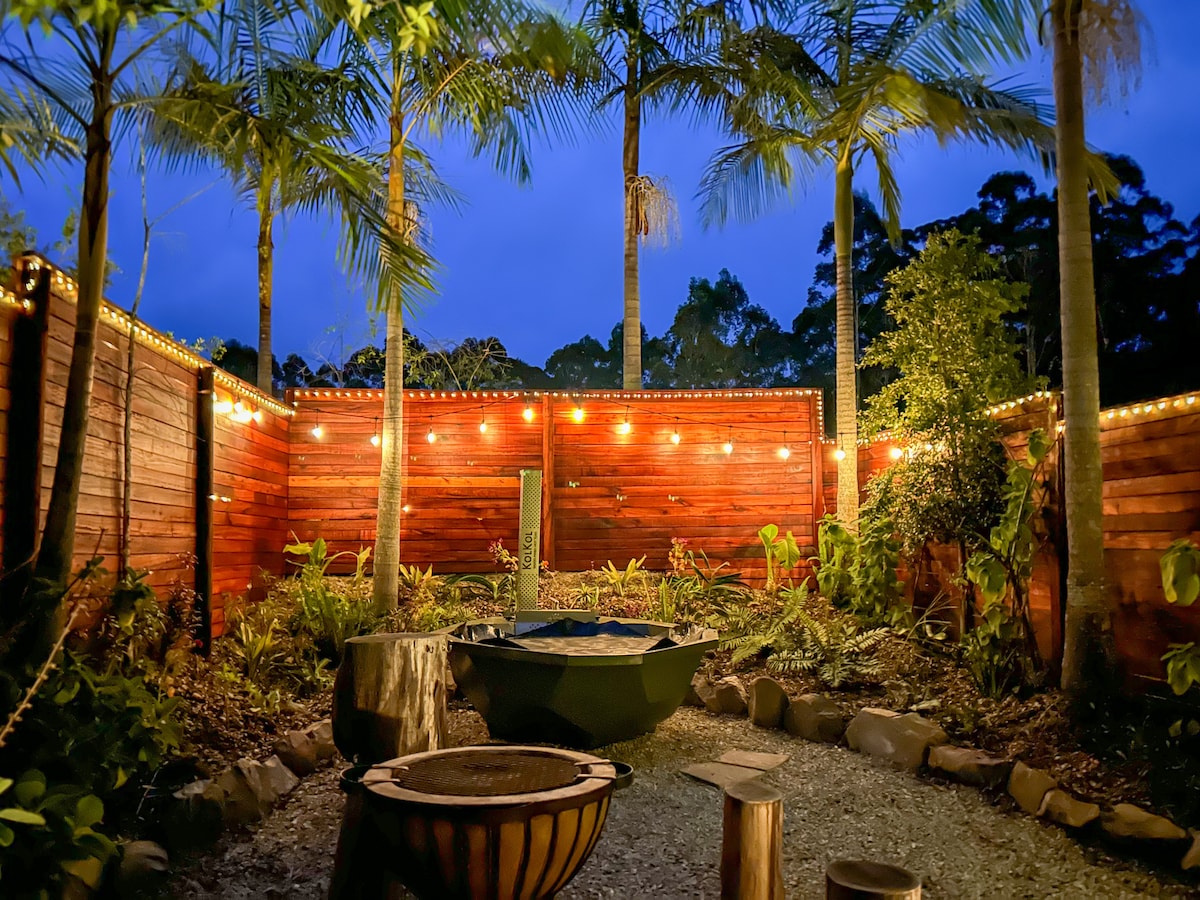 A private entertainment area is surrounded by lush greenery and illuminated by string lights. A fire pit and hot tub are nestled among natural stone features, creating an inviting outdoor setting. Tall palm trees rise above the wooden perimeter, enhancing the secluded atmosphere.