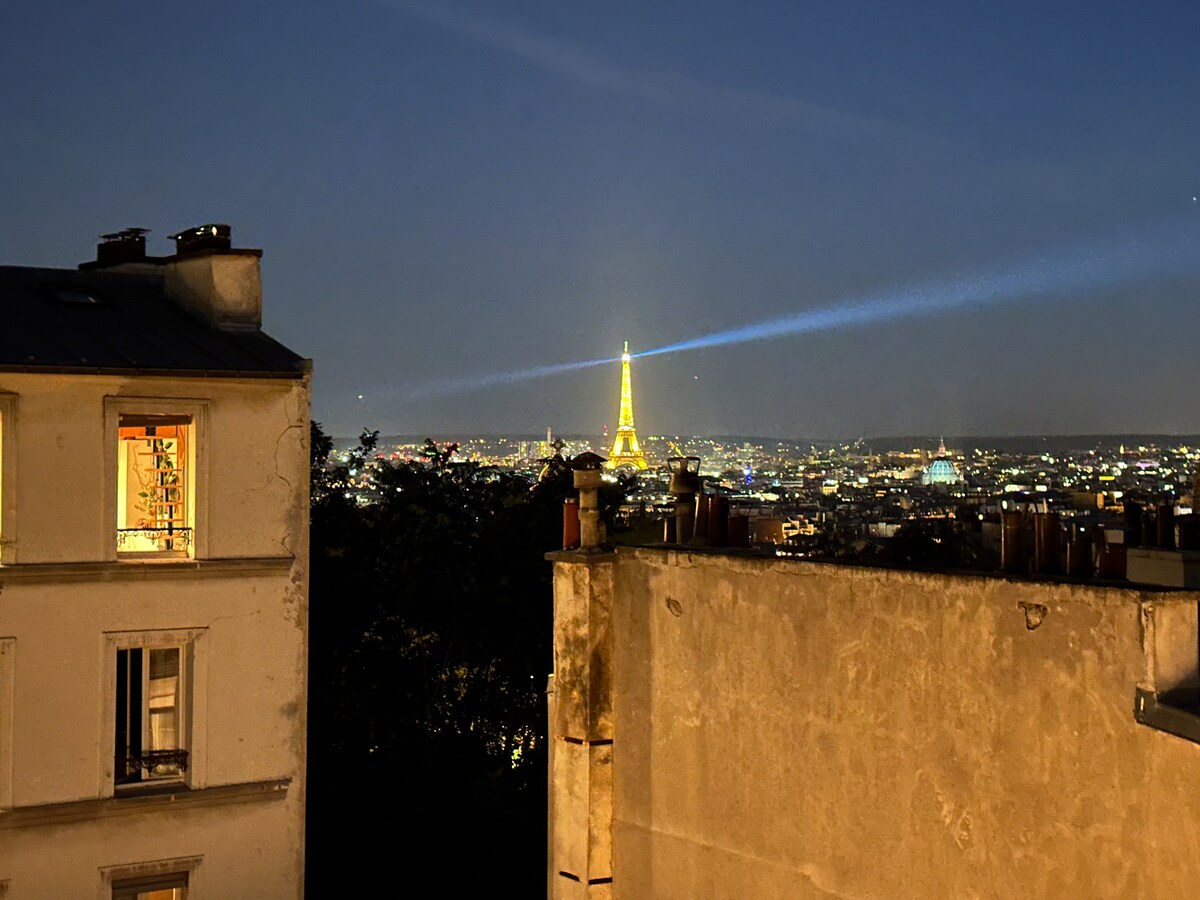A nighttime view reveals the Eiffel Tower illuminated against a dark sky, casting a warm glow across the cityscape. The foreground features the rustic rooftops of neighboring buildings, with a glimpse of a lit window showing a welcoming interior.