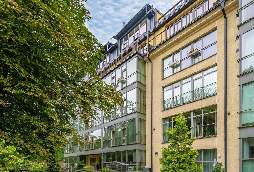 The exterior of a modern building is showcased, featuring large glass windows and balconies lined with greenery. Surrounding foliage adds a natural element, enhancing the building’s inviting presence in its tranquil setting.