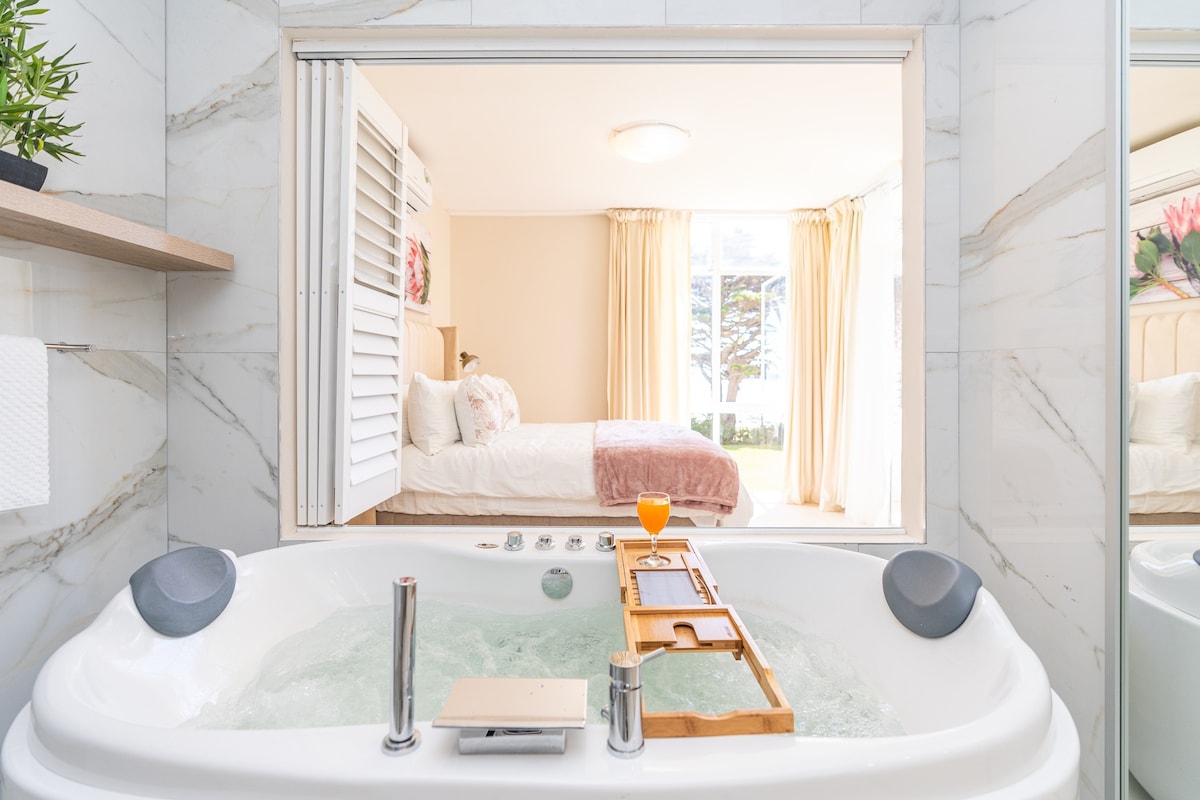 A well-appointed en-suite bathroom features a spacious jacuzzi tub, with a wooden tray holding a drink positioned in the center. The mirror reflects the adjoining bedroom, which is decorated in soft tones and framed by large windows that allow natural light to fill the space.