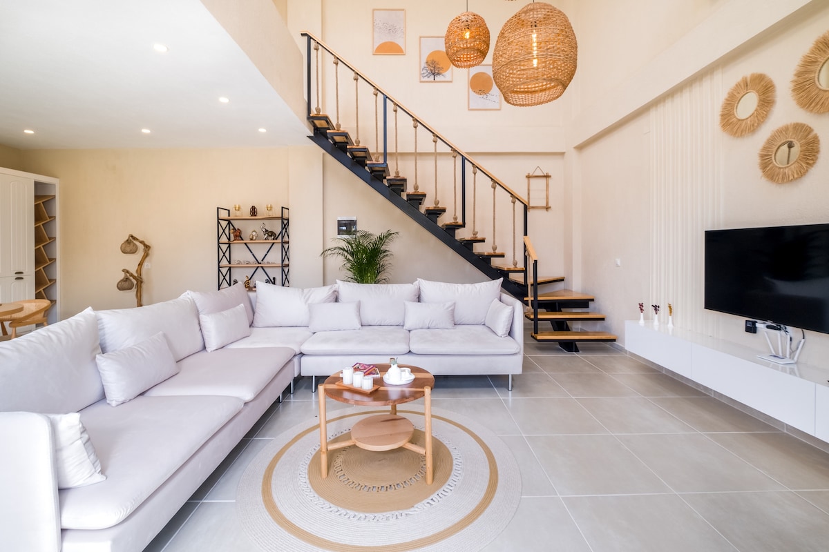 The living area features a soft, L-shaped sofa arranged around a simple wooden coffee table. Natural light fills the space, highlighting the neutral tones and textured wall decor. A staircase with elegant railings leads to the upper level, while a television is positioned against a white cabinetry unit.