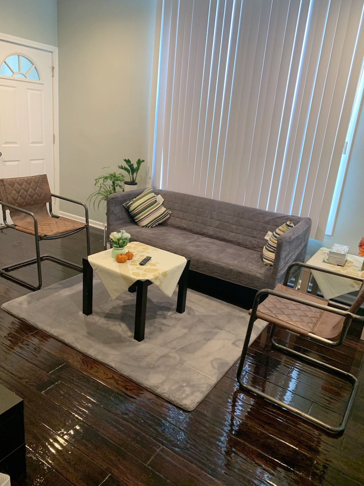 A cozy seating area features a gray sofa accompanied by two stylish chairs. A small table is centrally positioned, adorned with a decorative plant and a fruit centerpiece. Natural light filters through large vertical blinds, illuminating the polished wood floor.