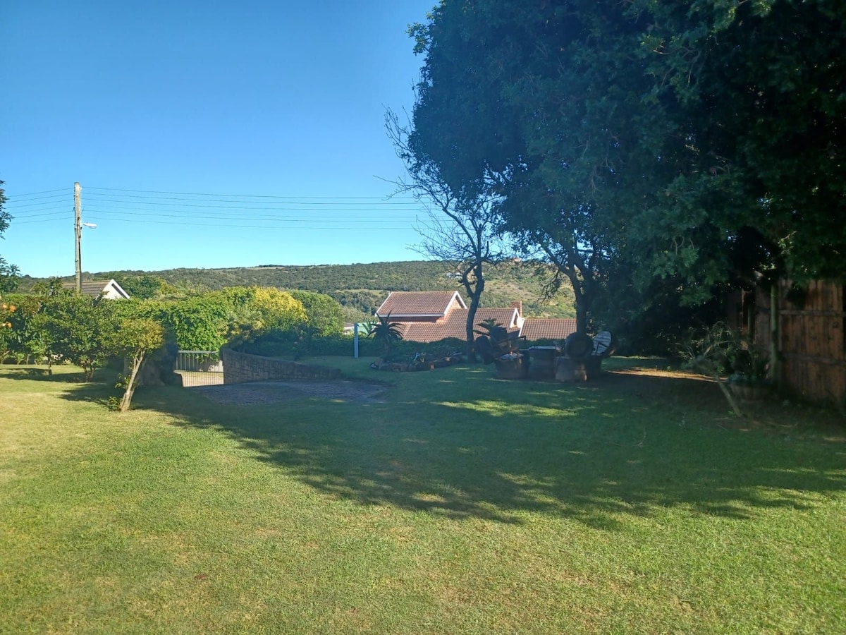 A tranquil garden area is surrounded by green grass and trees, providing a peaceful atmosphere. A pathway leads through the space, with glimpses of nearby cottages visible in the background. The landscape is complemented by the distant hills, enhancing a sense of serenity.