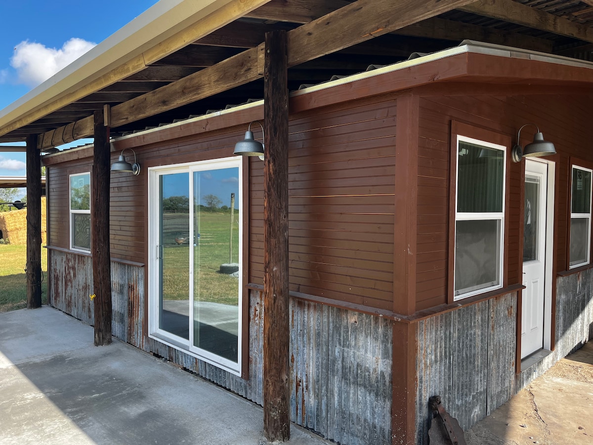 The exterior of the cabin is shown, featuring a combination of wooden and metal paneling. Large glass doors provide access to the interior, framed by simple light fixtures. The surrounding landscape is open and grassy, with blue sky visible above.