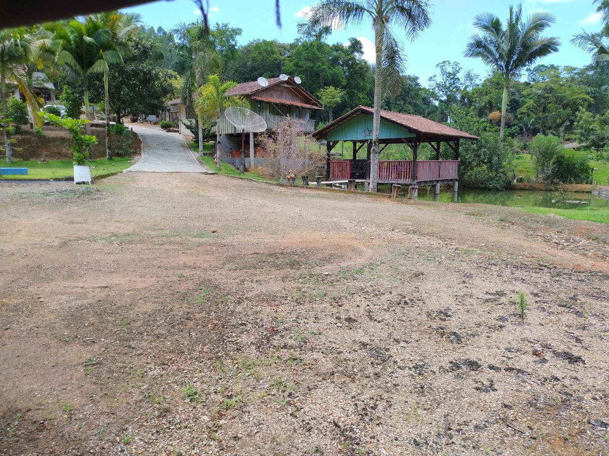 A gravel path is flanked by lush greenery, leading to distinct wooden structures. One structure features a covered porch, while another has an open design with a sloped roof. Tropical palm trees add to the serene setting, creating a peaceful atmosphere.