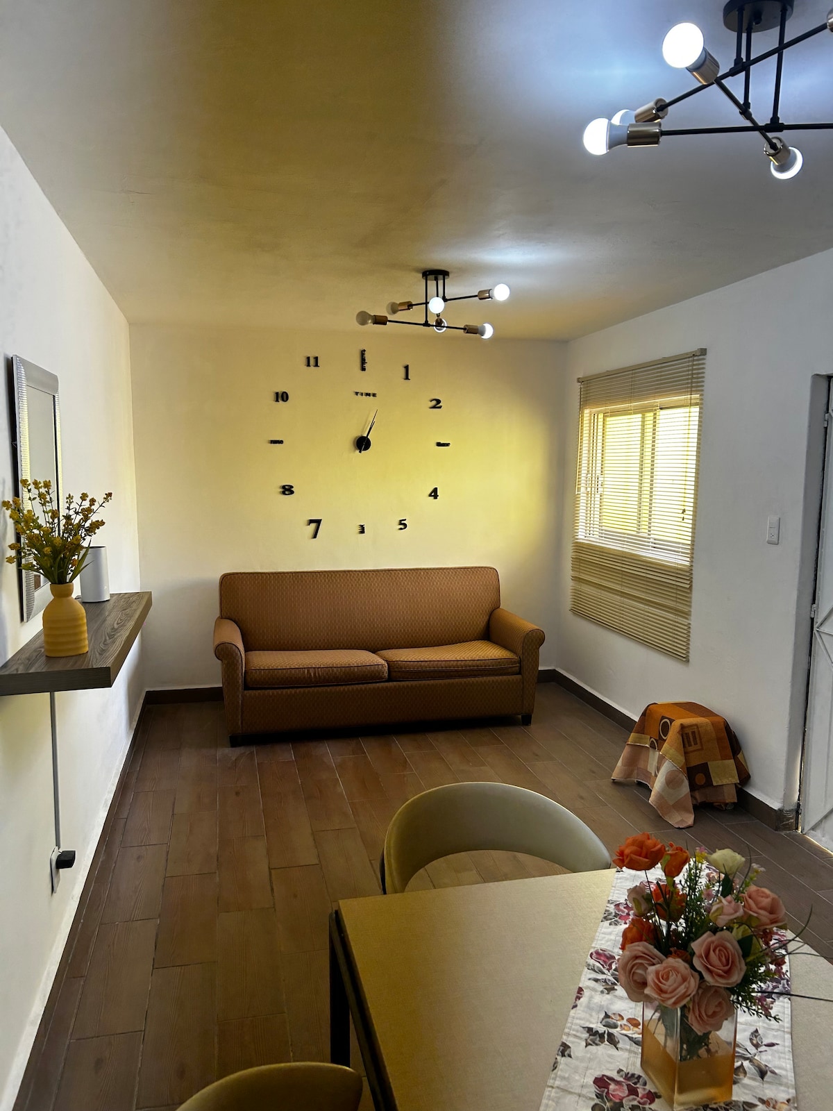 A cozy living area features a brown sofa facing a charming wall clock, with natural light filtering in through a window covered by horizontal blinds. A dining table with floral decor is present, complemented by round chairs and a light fixture with multiple bulbs above.