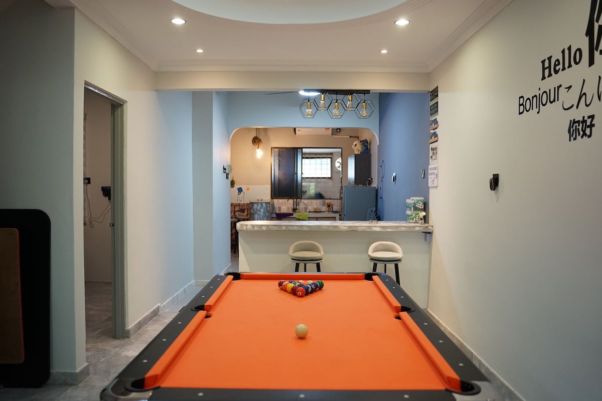 A vibrant game room is presented, featuring an orange pool table at the forefront. Soft lighting is visible throughout the space, with a kitchenette in the background. Wall decorations in multiple languages offer a welcoming touch, enhancing the area's inviting atmosphere.