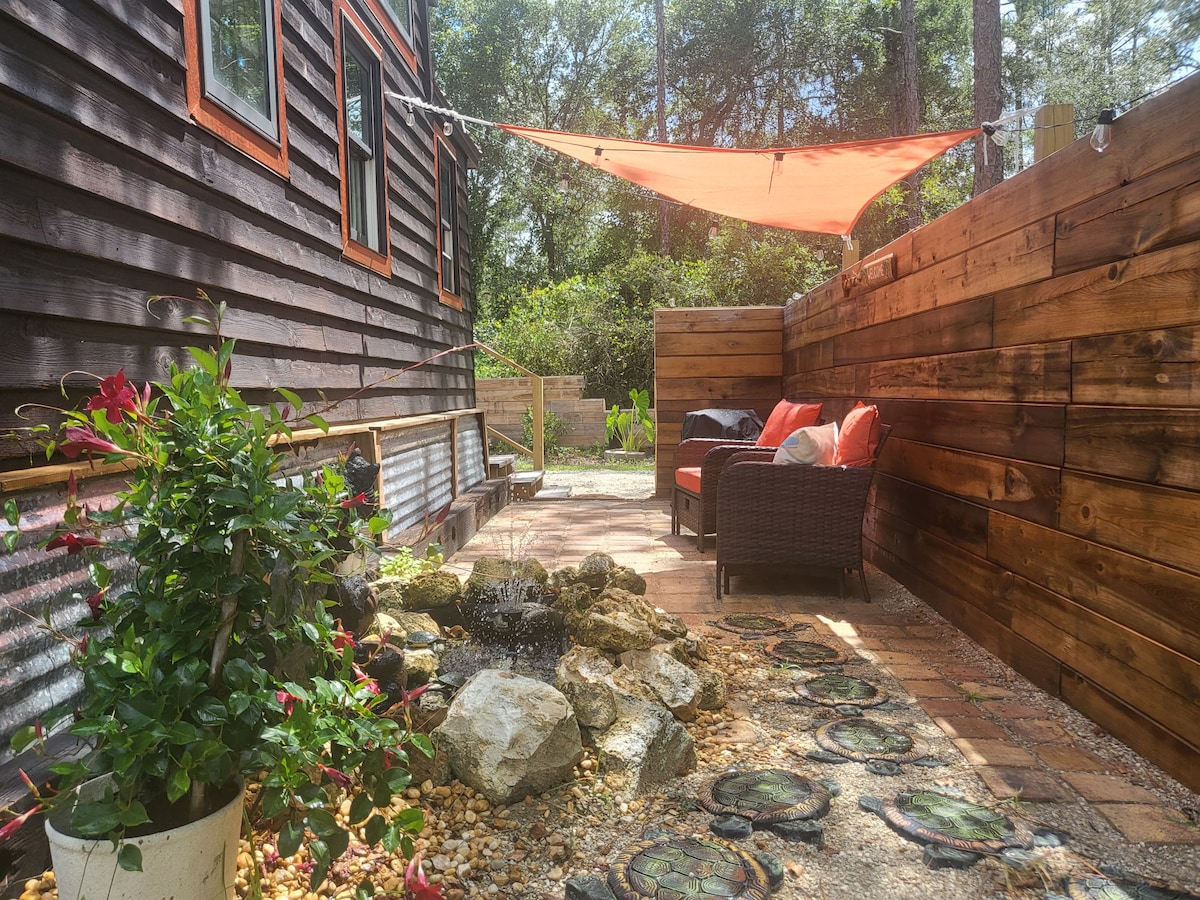 A small patio area features a decorative fountain surrounded by stones and gravel. Two comfortable chairs sit beneath a vibrant orange shade sail, while lush greenery is visible in the background. A wooden fence encloses the space, providing a sense of privacy.