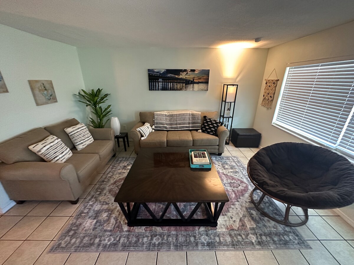 The living area features two comfortable sofas and a large, round chair, situated around a contemporary coffee table. A decorative area rug covers the tile floor, and natural light filters through the blinds on the windows. A small plant adds a touch of greenery to the space.
