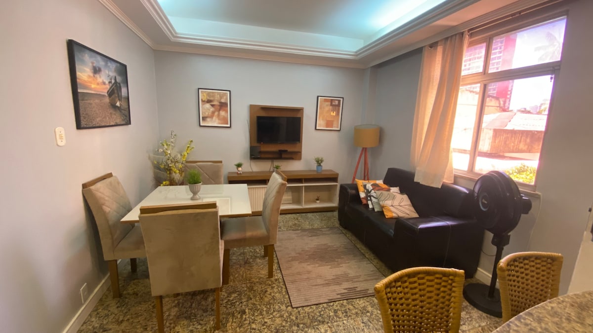 A cozy living area features a dark sofa and a light, modern dining table surrounded by four chairs. Two framed artworks are displayed on the wall, and a television is mounted on a wooden console. Natural light enters through the large window, adorned with light curtains.