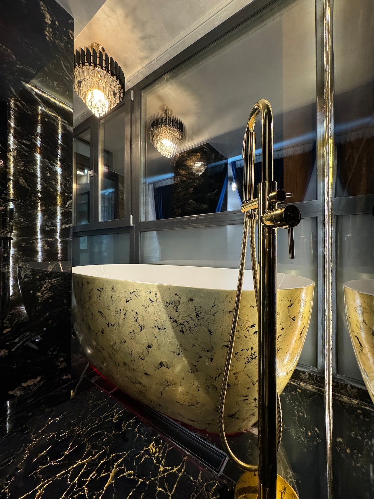 A modern bathroom features a stylish freestanding tub with a gold-patterned exterior, complemented by a sleek gold faucet. Large windows allow natural light to filter through, and an elegant chandelier provides ambient lighting, enhancing the luxurious setting.