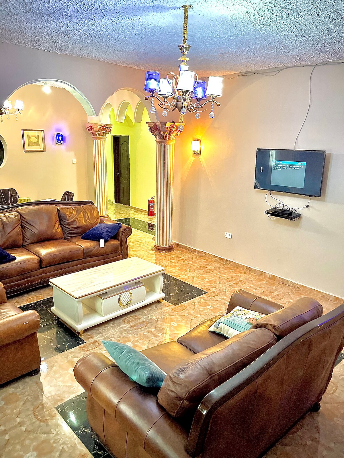 A spacious living area is presented, featuring comfortable leather sofas arranged around a light-colored coffee table. Decorative columns lead to adjacent rooms, while a modern chandelier provides soft lighting. A wall-mounted television is positioned for easy viewing.