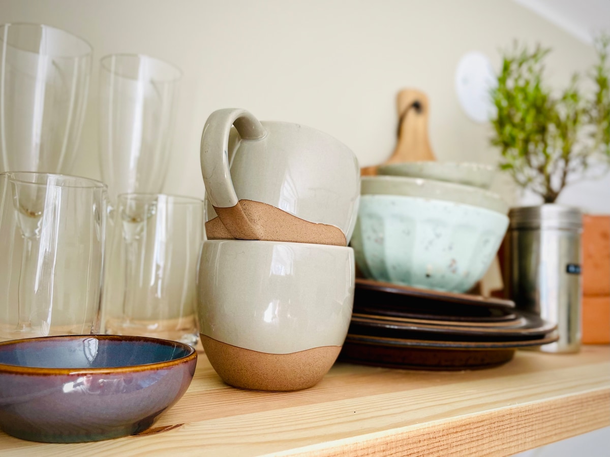 A collection of dishware is displayed on a wooden shelf, featuring various items such as stacked cups, drinking glasses, and bowls. A small plant adds a touch of greenery, while utensils and serving dishes are neatly arranged, creating a functional kitchen space.