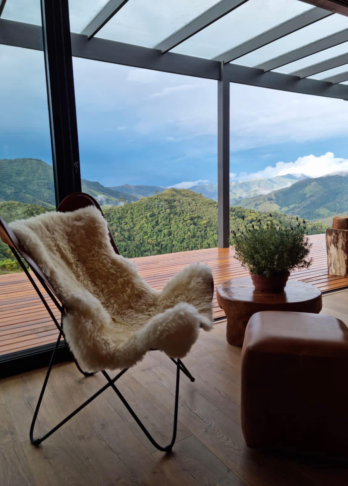 A cozy seating area features a soft, textured chair beside a rustic wooden table. Large glass doors frame the expansive mountain views, inviting natural light into the space. A potted plant adds a touch of greenery, enhancing the serene atmosphere.