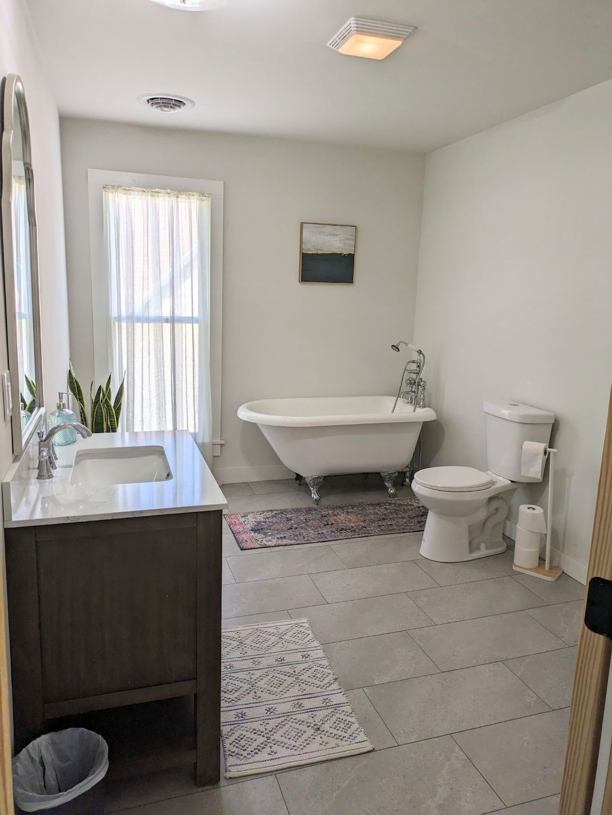A bright bathroom features a freestanding clawfoot tub and a modern vanity with a sink. A window with sheer curtains allows natural light to enter. The space includes a toilet and decorative rugs on the tiled floor, enhancing the room's functionality.