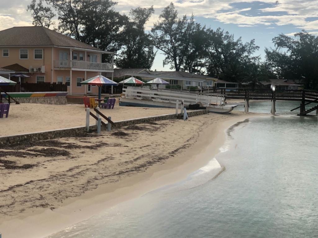 A sandy beach area is framed by a calm blue waterway. Colorful umbrellas provide shade over seating arrangements on the sand. In the background, a two-story villa is visible alongside quaint cottages, with lush greenery rising behind them.