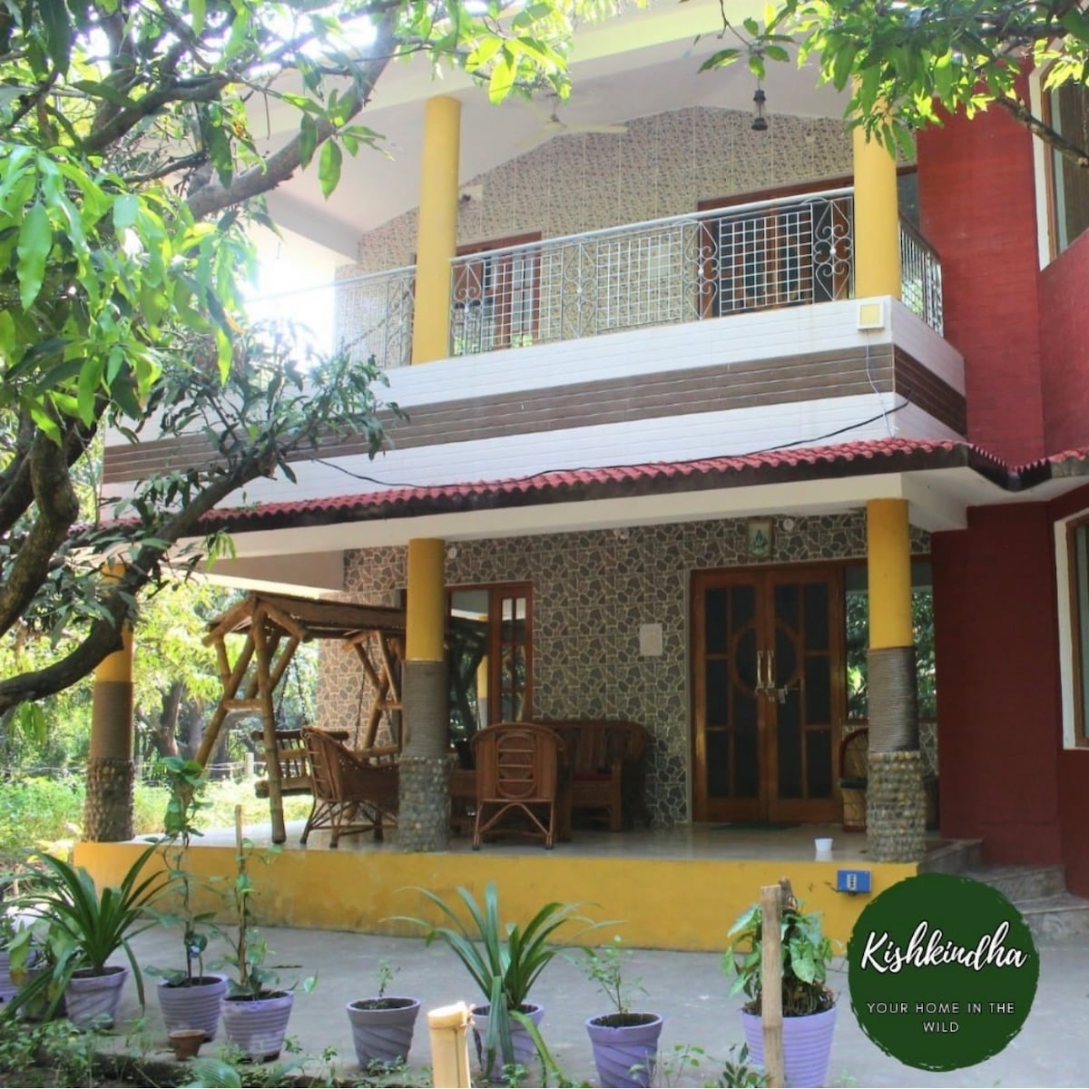 The two-story house is surrounded by greenery, featuring a welcoming porch with wooden rocking chairs. Bright yellow columns support the balcony above, while patterned stone walls provide texture. Potted plants are arranged along the base, enhancing the natural setting.