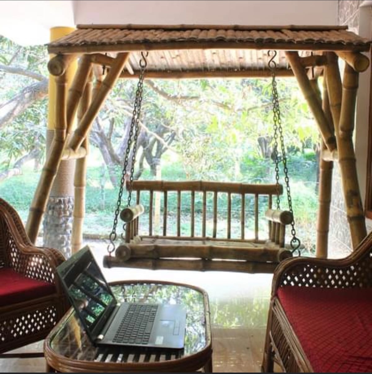 A bamboo swing is positioned within a bright area, surrounded by greenery. Two woven chairs with red cushions are placed nearby, and a laptop rests on a glass-top table, creating a comfortable workspace alongside a relaxing seating option.