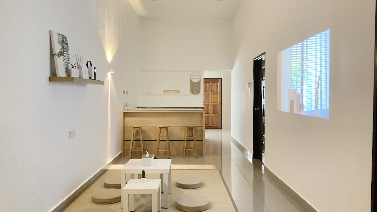 An open living space features a light-colored wooden bar with three stools positioned in front. Two low coffee tables and circular cushions are arranged on a light rug, while a wall displays a projection of colorful graphics. Natural light illuminates the area.