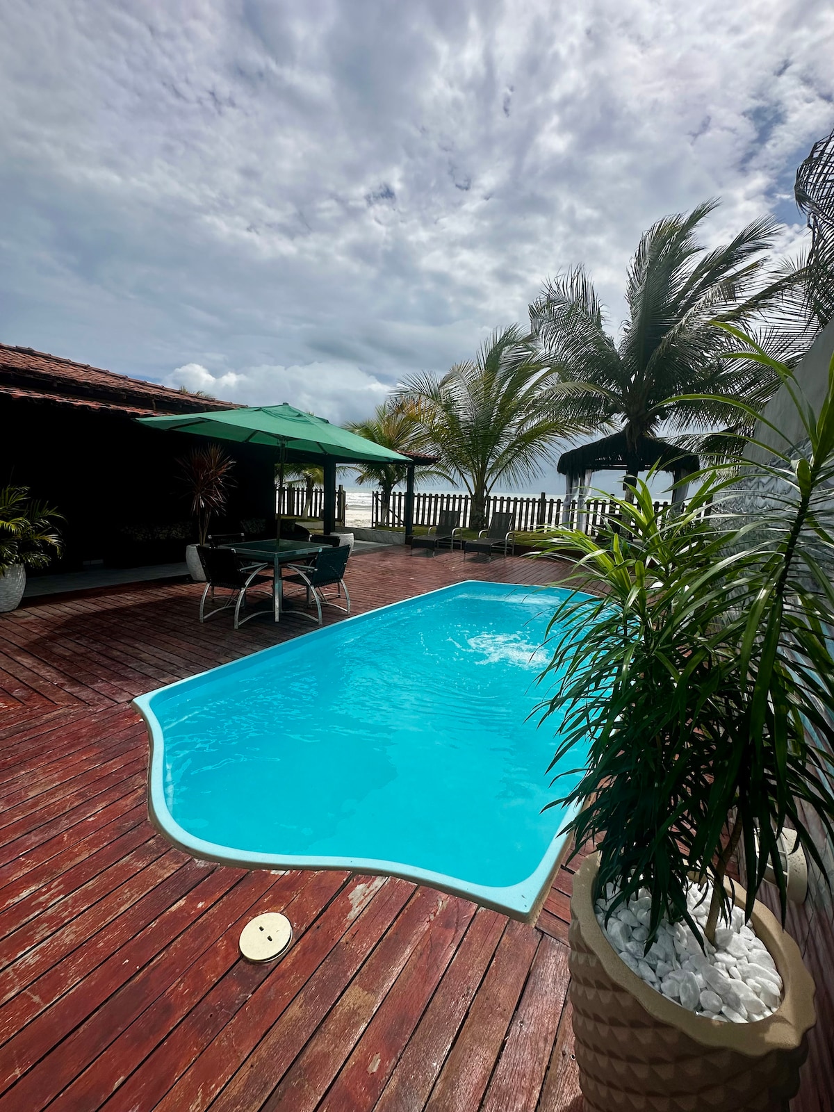 A private pool with clear blue water occupies a wooden deck surrounded by tropical greenery. Comfortable seating under a green umbrella enhances the outdoor space, while palm trees sway in the gentle breeze nearby, creating a relaxing environment for guests.