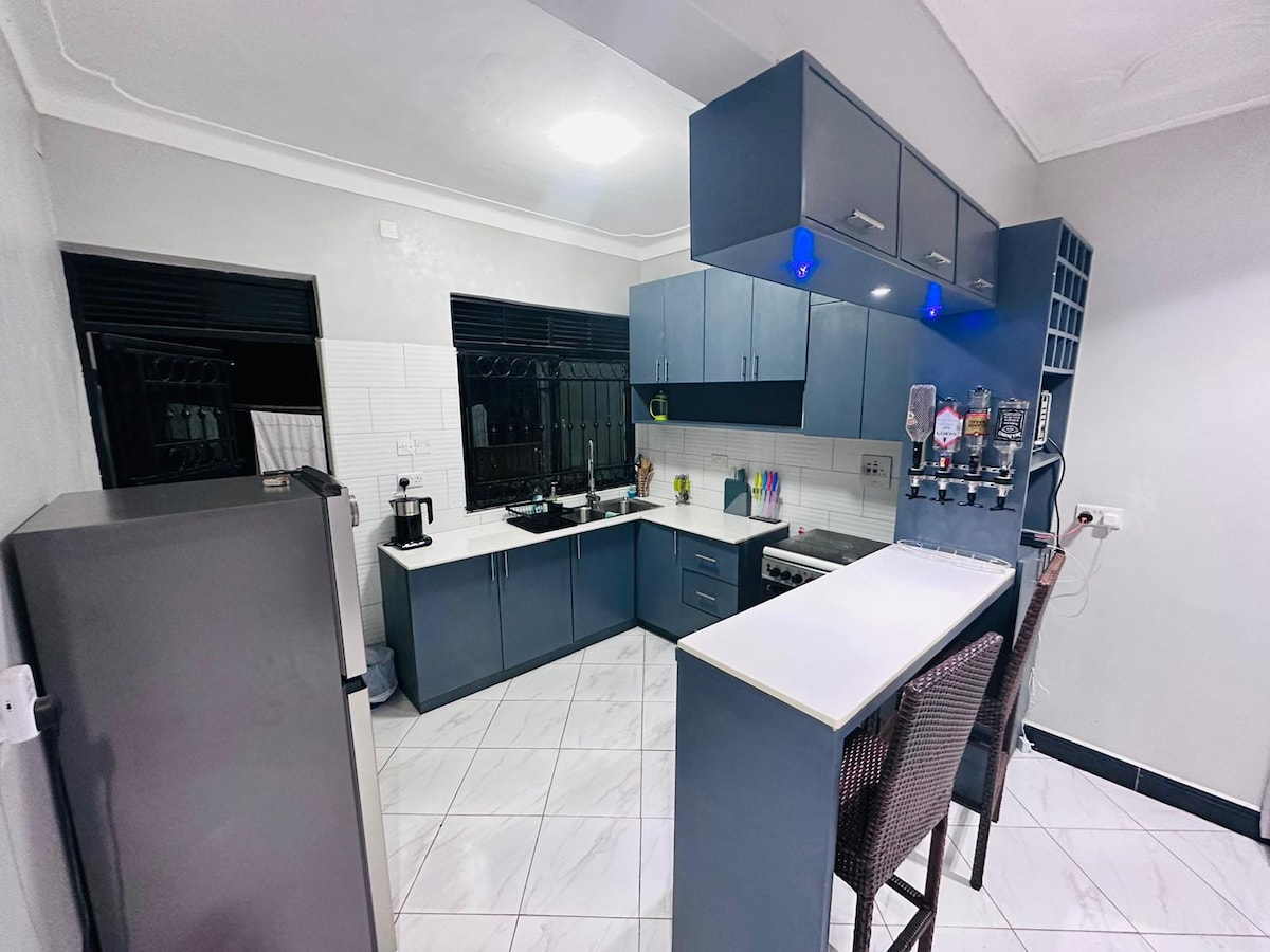 The kitchen showcases modern cabinetry in a deep blue hue, complemented by white countertops. Appliances include a refrigerator and a stove. A bar area with seating for two enhances functionality, while ample natural light filters through two windows, creating a bright cooking environment.