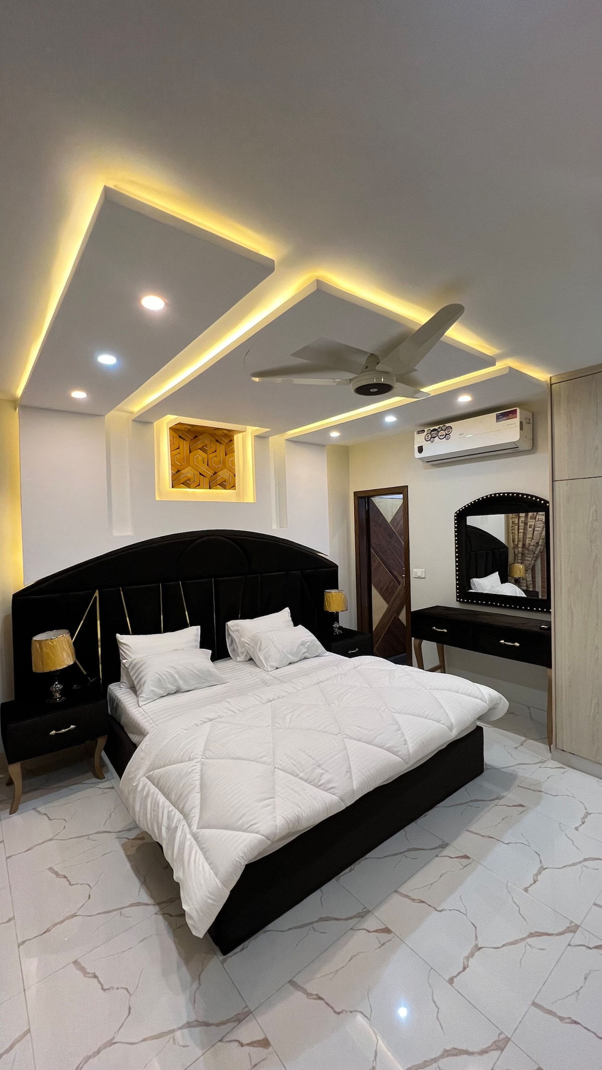 A modern sleeping area features a stylish black bed with a white quilt, flanked by two bedside tables with lamps. The ceiling is outlined with soft lighting, and a fan is mounted above. A decorative wall element adds interest, while the flooring is a sleek tile pattern.