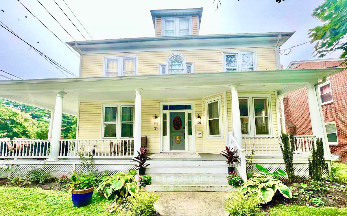 A charming yellow house stands with a welcoming porch framed by tall columns. Lush greenery surrounds the entrance, including varied foliage and flower beds. Large windows allow natural light to illuminate the inviting exterior, while decorative elements enhance the home’s character.