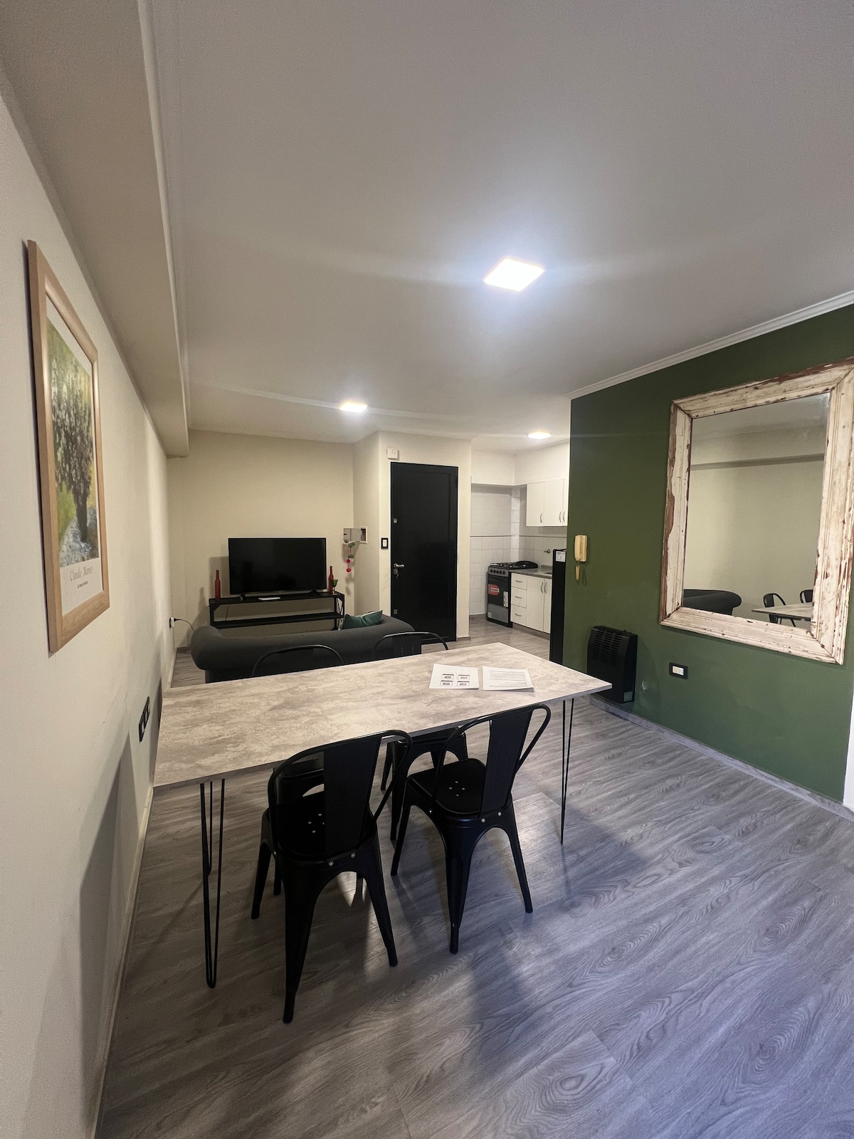 The living area features a dining table with four black chairs, positioned against a green accent wall. A comfortable sofa and a television are visible, while a large mirror adds depth to the space. A kitchenette is subtly integrated in the background.