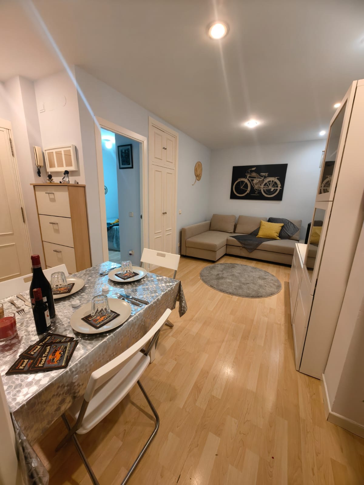 A light-filled dining area features a table set for four, complemented by white chairs. A cozy seating area with a sofa and decorative cushions is positioned nearby. A bicycle artwork adorns the wall, while warm lighting creates an inviting ambiance throughout the space.