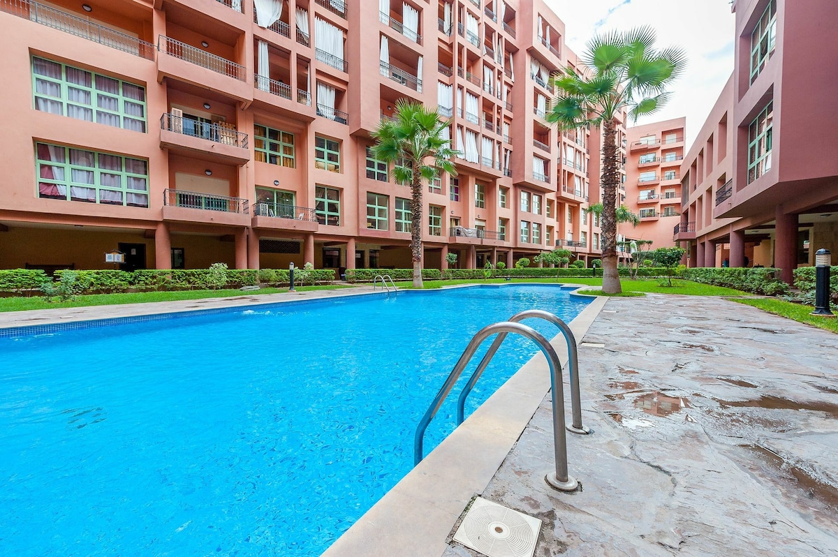 A clear view of the swimming pool is presented, featuring a vibrant blue surface surrounded by landscaped grounds. Palm trees line the pool area, while the orange-red hues of the residential building are visible in the background, contributing to a relaxed atmosphere.
