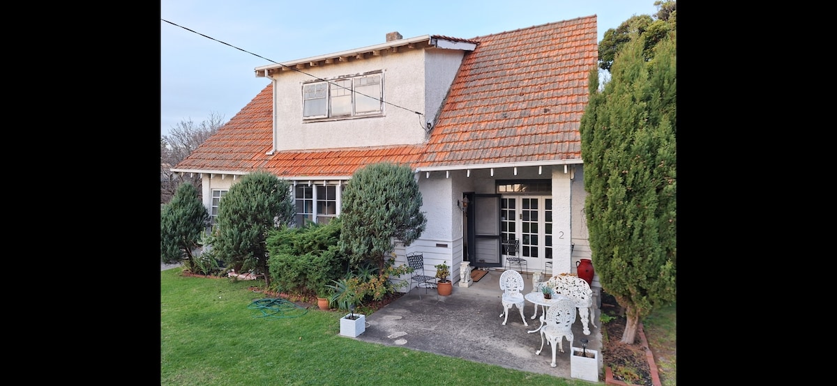A charming two-story cottage features a red-tiled roof and well-maintained gardens. Outdoor seating includes white, intricate chairs and a small table, situated on a stone patio. Lush greenery surrounds the property, while large windows enhance the home’s inviting appearance.