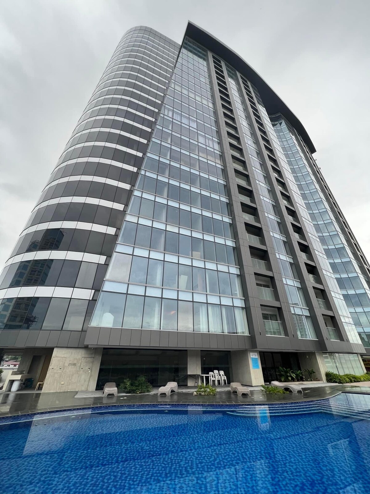 The modern high-rise building features numerous large glass windows, reflecting the sky above. A pool with a vibrant blue surface sits at the foreground, framed by greenery and outdoor seating options. The sleek design emphasizes an urban lifestyle in the Abreeza District.