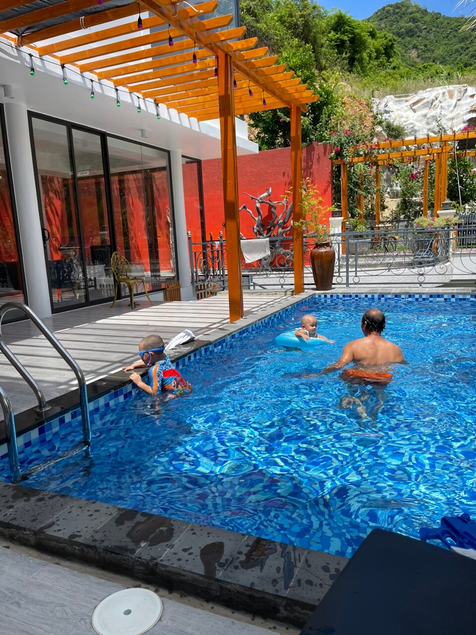 A swimming pool is surrounded by vibrant greenery and colorful landscaping, with sunlight reflecting off its clear blue water. Two children are playing in the shallow end, while an adult relaxes nearby on the pool steps. Decorative lights hang from a wooden pergola overhead.
